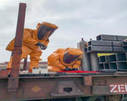 Two individuals in yellow hazardous materials suits inspect a simulated hazardous device during a training drill.