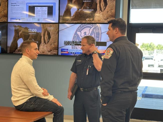 Three men talking. One sitting and two standing. 