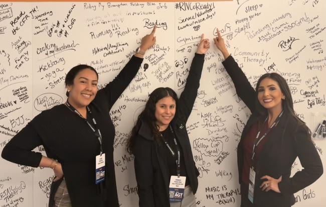 Three women standing and pointing to a wall with a lot of writing. / Tres mujeres de pie y señalando una pared con mucha escritura. 