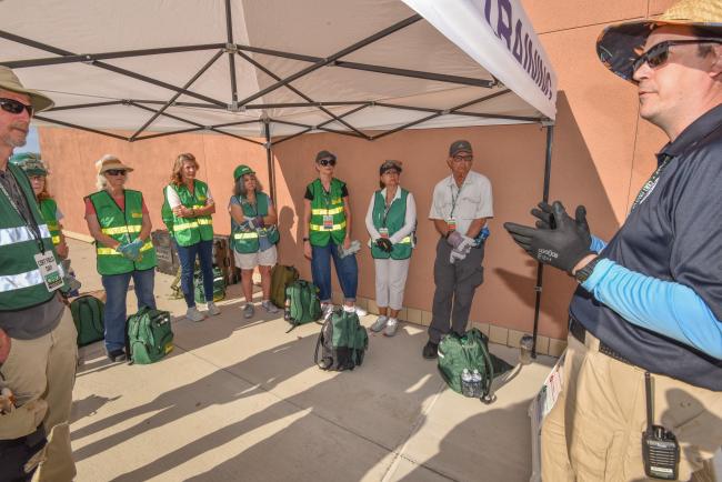 A group of people wearing green CERT gear listen to an instructor under a tent.