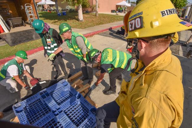 A firefighter looks on as CERT members use a leverage tool to lift a heavy object.