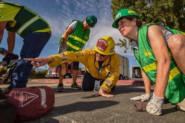 A firefighter in firefighting gear points to give instruction to a woman in CERT gear.