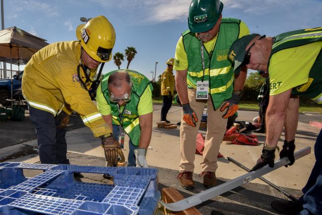 A Firefighter gives direction to a group of people  in CERT uniforms with heavy equipment nearby.